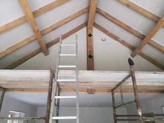 Plasterboard fitted around exposed timber beams at a gable, with a ladder and scaffolding below.