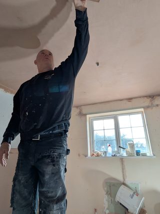 Plasterer reaching up with a trowel to skim a ceiling above unfinished walls.