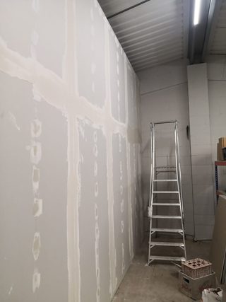 Long plasterboard wall with compound over joints and fixings, beside a stepladder.