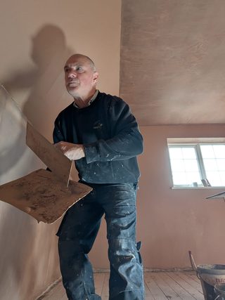 Plasterer holding a hawk and trowel beside a freshly plastered wall.