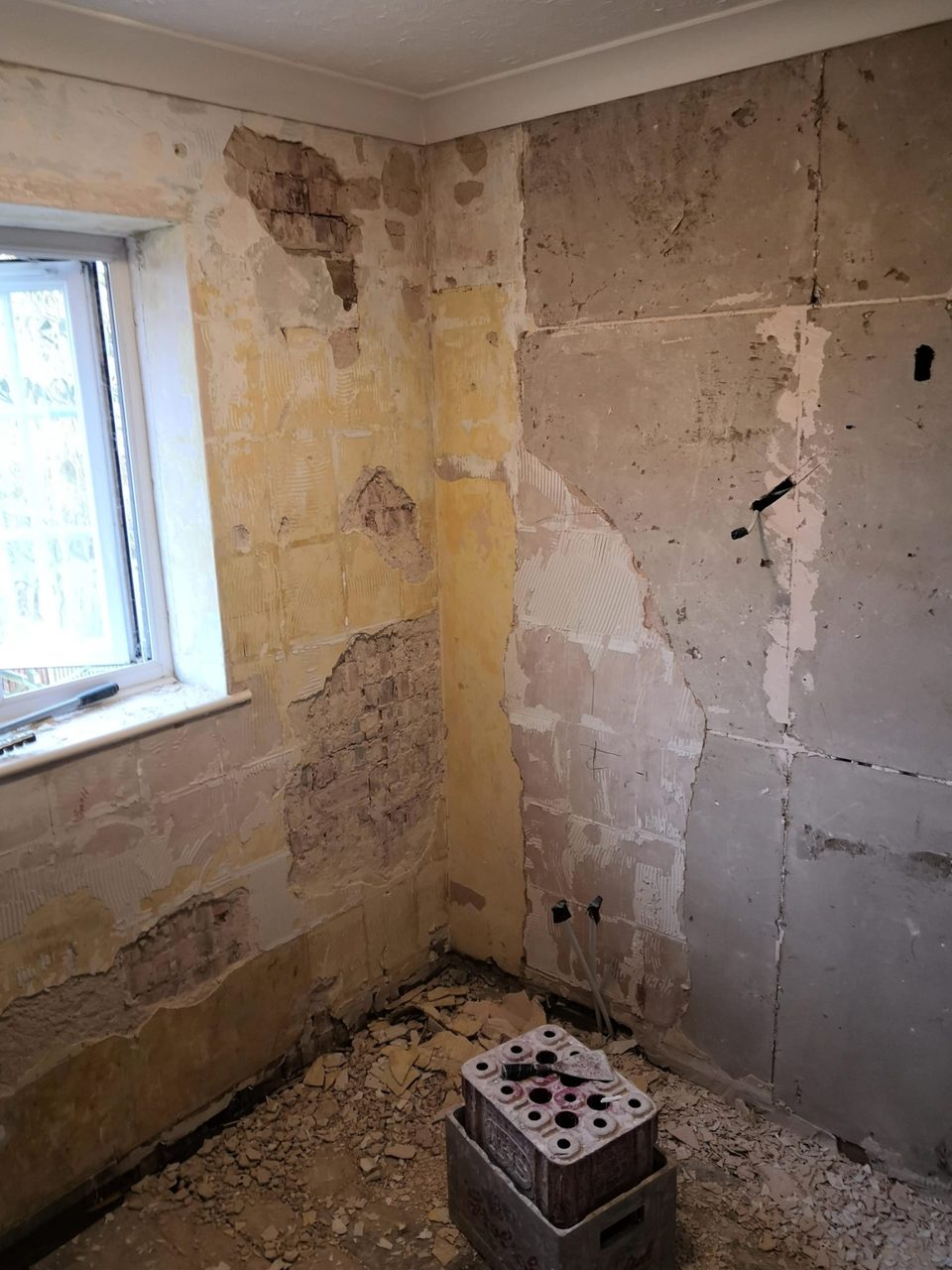Stripped corner beside a frosted window with damaged plaster, adhesive marks and pipe outlets.