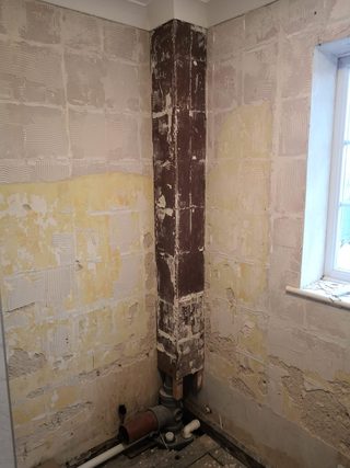 Stripped walls and pipe boxing beside a frosted window, with exposed adhesive marks and pipework.