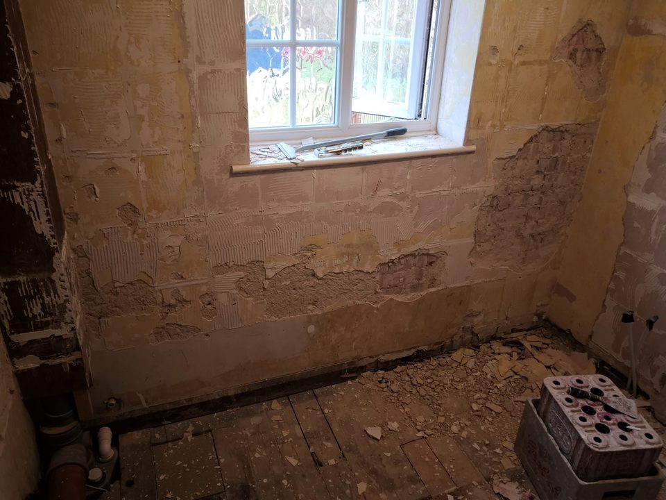 Stripped wall beneath a frosted window with adhesive marks and exposed patches of brickwork.